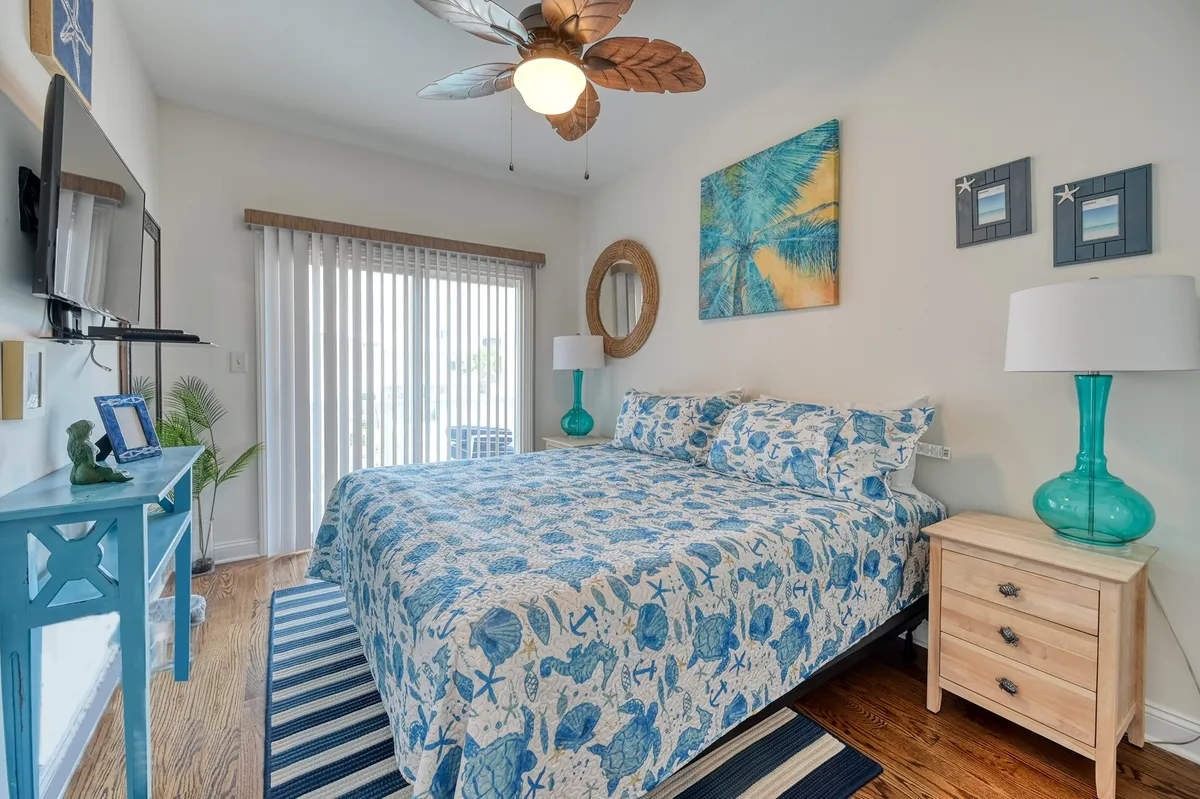 A cozy bedroom with a plush bed, decorative wall art, ceiling fan, nightstand, and a television, bathed in warm sunlight filtering through the curtains. The room has a relaxed, beachy vibe and feels perfect for unwinding after a busy day.