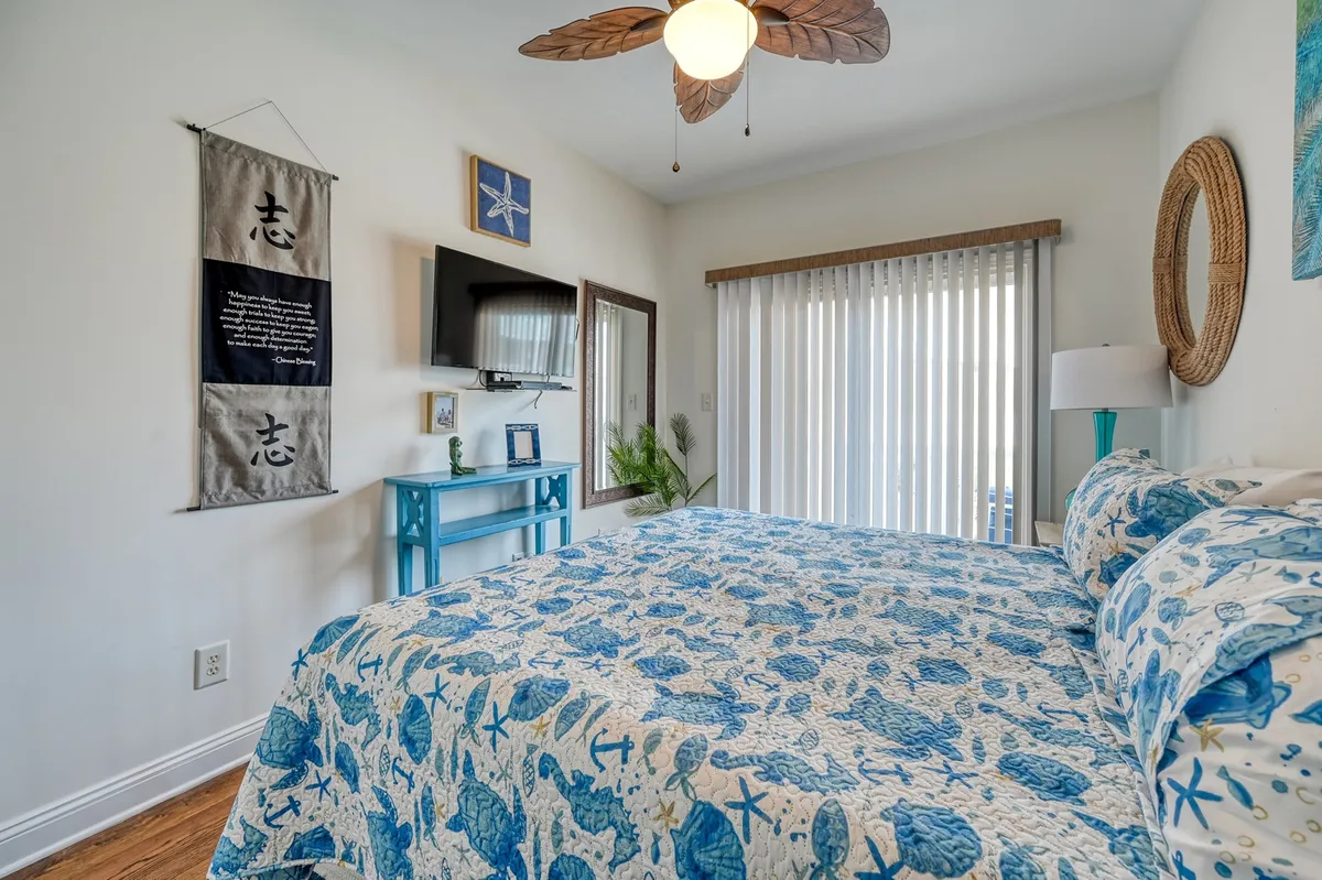 A tranquil bedroom filled with natural light, featuring blue and white patterned bedding, beach-themed decor, a paddle ceiling fan, and a large window with blinds that invites the outside in. The calm, beachy ambiance evokes restful mornings and a peaceful retreat.