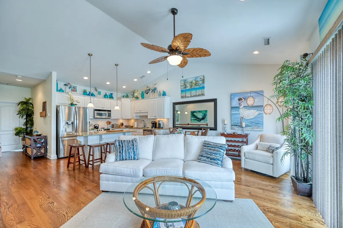 A comfortable, airy living room with a plush white sofa, decorative ceiling fan, and modern coffee table. Large windows suggest a connection to the outdoors and provide ample natural light, creating a cozy yet elegant social space.