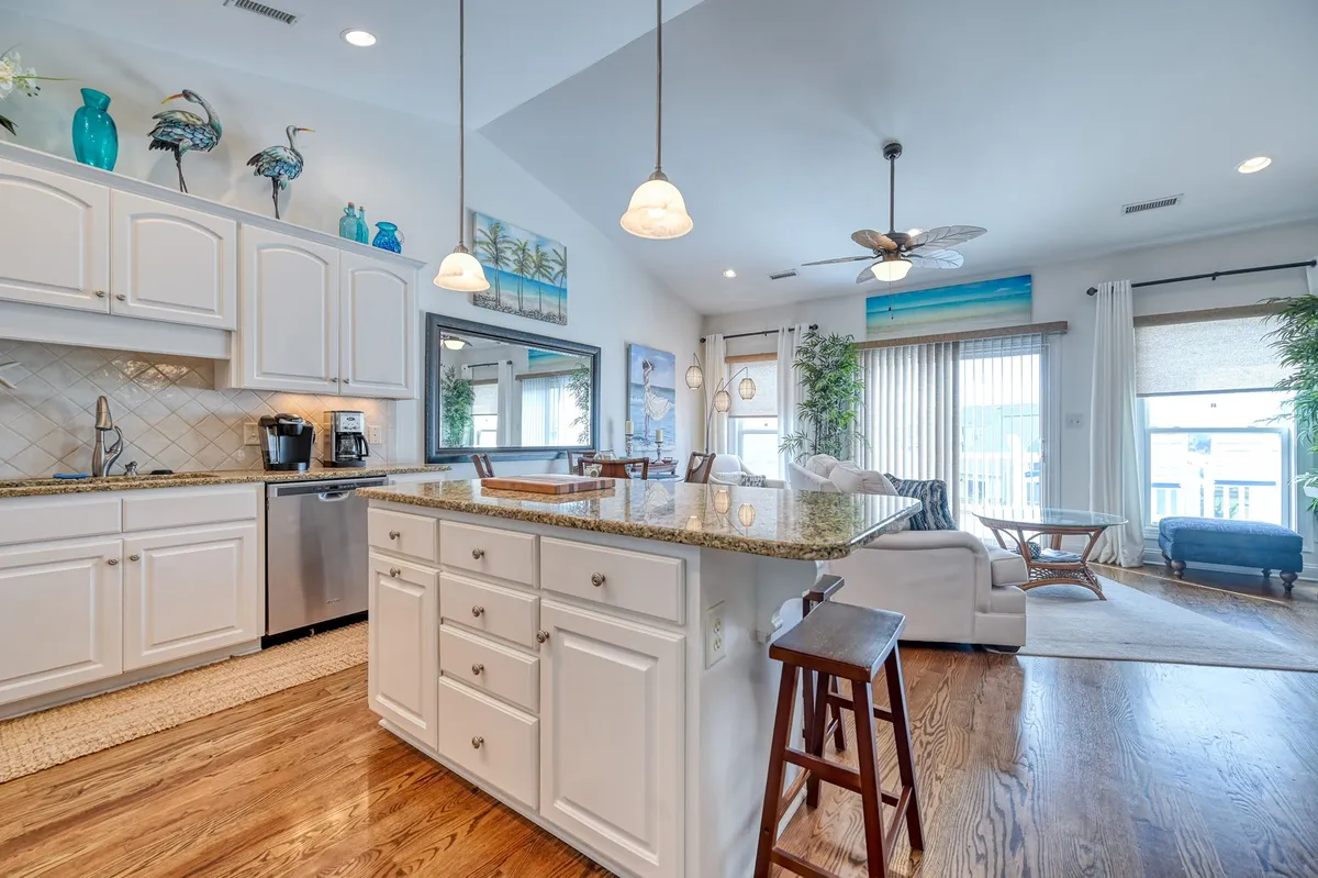 The image showcases a bright and inviting kitchen with a sleek granite countertop and a spacious kitchen island, surrounded by tasteful decorative wall art. Large windows flood the space with natural light, creating a cheerful and welcoming atmosphere ideal for cooking, entertaining, and gathering with family or friends.