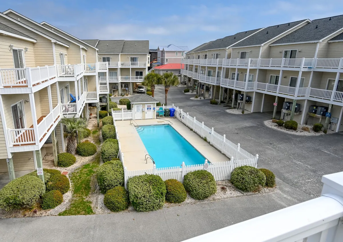 A bright, inviting exterior shot of a swimming pool bordered by green shrubbery, with balconies of the surrounding apartments in the background. The poolside atmosphere feels lively yet relaxed, ideal for socializing or taking a dip.