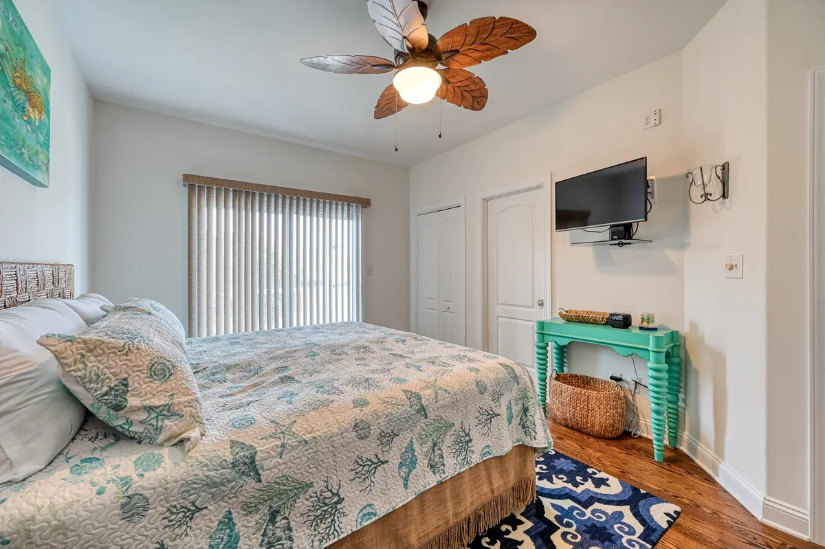 A serene bedroom with a patterned quilted bed, a ceiling fan designed like palm leaves, decorative coastal wall art, and a green accent table. The room offers a calming, nature-inspired vibe, ideal for unwinding after a day of exploration.