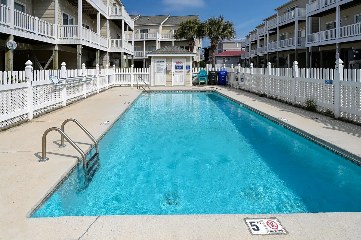 A bright community pool area surrounded by a white fence and poolside steps, complemented by a nearby lifeguard station. The atmosphere is relaxed and social, with ample space for swimming and sunbathing.