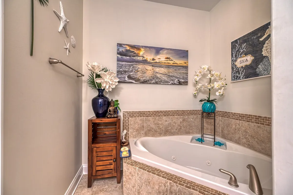 This image features a whirlpool garden jetted tub set beneath ocean-themed artwork, complemented by a wooden side table with decorative items and a vase of flowers. The space is bathed in soft, soothing light, enhancing the spa-like atmosphere.