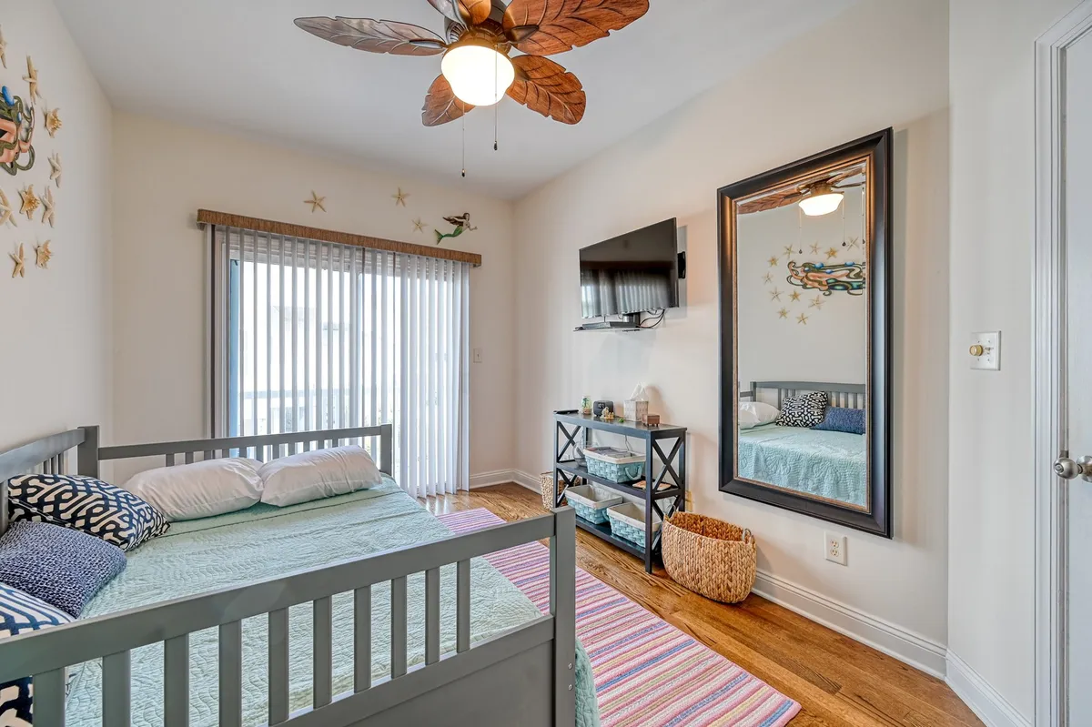 This image shows the primary king suite, featuring a bed with stylish patterned bedding, decorative wall art, a leaf-shaped ceiling fan, a mirrored decorative frame, and a wooden TV stand. Natural light fills the room, evoking a sense of comfort and tranquility, perfectly aligning with the concept of a restful private paradise.