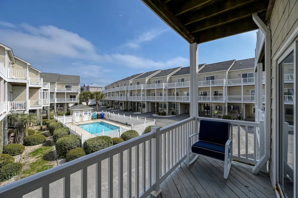 The image captures a tranquil view of the swimming pool from a balcony seating area, with landscaped bushes and the building's architecture in the background. The peaceful setting exemplifies the inviting pool views available from the suite's balcony.